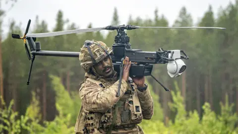 PA Media A British soldier in battle fatigues carries a a single-rotor helicopter style Ghost drone on his shoulder in a forest