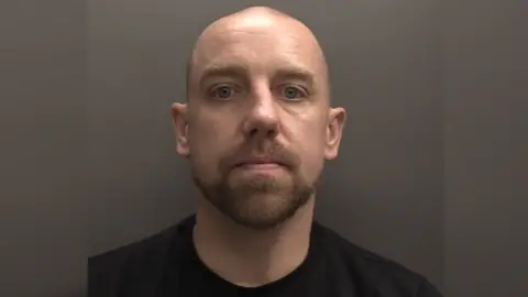 A police mugshot of Thomas Michael Sanigar, who is facing the camera. He is bald and has a short brown moustache and beard. He is wearing a black T-shirt. 