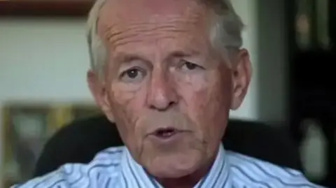 John Smyth looks on during a court hearing. He's sat on a chair and wearing a stripy shirt.
