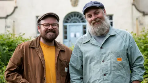 Shelley Richmond/Hello Halo/BBC Scotland Banjo Beale and his husband Ro stand in front of the derelict mansion Ulva House, a grand but shabby building - its pale blue front door visible behind them. Banjo, with his trademark beard, glasses and flat cap, smiles alongside Ro, also in a cap, who has a greying long beard.