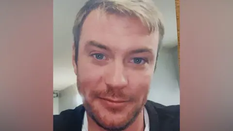 Sussex Police A blond man with blue eyes, with a light stubble, and wearing a black jacket. 