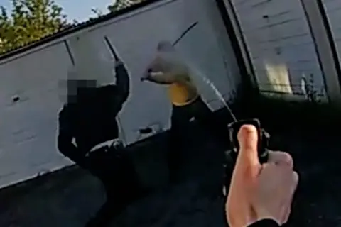 A police officer sprays a substance towards a man wielding a sword near another police officer, whose baton is raised