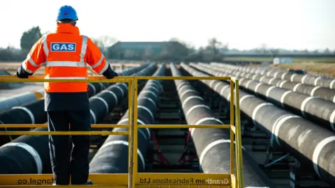 A man wearing an orange hi-vis jacket standing with his back to the camera on a platform, looking at pipelines, which aim to deliver clean hydrogen.