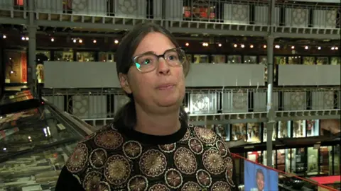 Laura Van Broekhoven from the shoulders up. She has medium length brown hair and glasses. She is stood in the pitt rivers, and behind her there are various display cases.