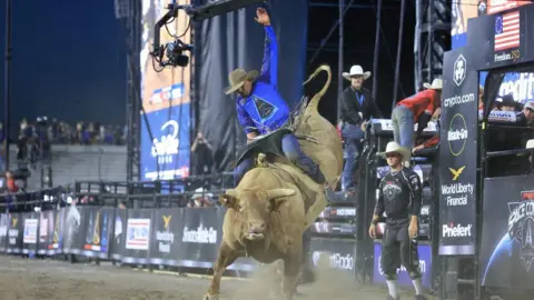 A professional bull rider, wearing a cowboy hat and blue shirt featuring the triangular logo of US Space Force, clings on while riding on the back of a massive, brown horned bull in a stadium filled with dirt 