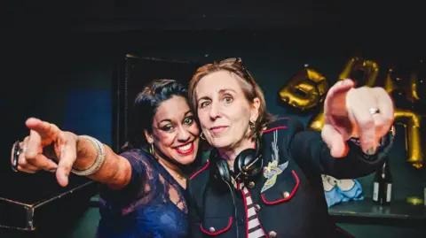 Two women - Aarti Joshi and Kirsty Wark - standing behind the decks in a club. Both are pointing towards the camera, with headphones round the neck of Kirsty Wark.