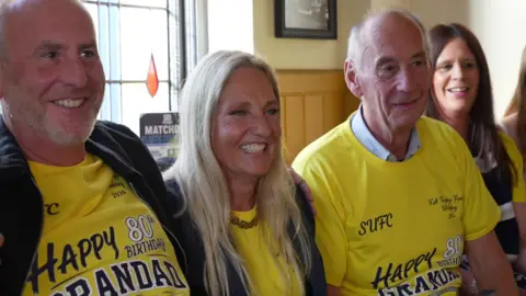 Julian, Vivienne and Doug Wantling at a pub. They are all smiling and wearing yellow, Southend-themed T-shirts that say "happy 80th birthday grandad".