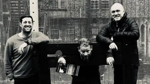 Sussex Trail Events Black and white photo of Chris, Jay and Danny at Shepton Mallet Prison. All three are laughing while Jay has his head and hands through a set of stocks.