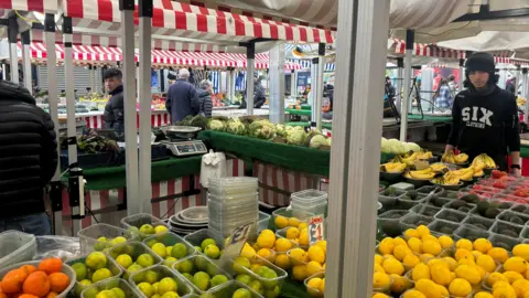 A colourful market stall packed with citrus and other fruit and veg under red and white awnings.