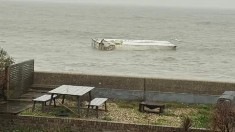A container floating in the sea. A garden with table and benches is in the foreground.