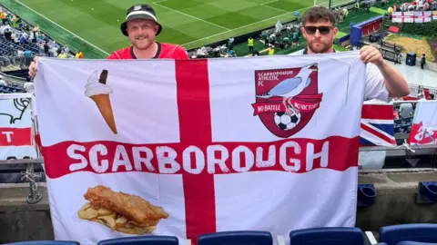 Two men holding an England flag with Scarborough written on it. The flag features the crest of Scarborough Athletic FC, as well as an ice cream and a portion of fish and chips.