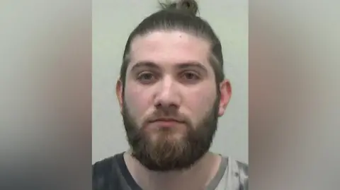 Northumbria Police photo of Hyseni. He has long black hair tied in a bun and a brown beard.