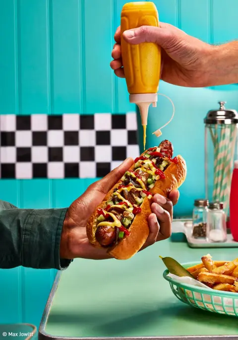 Max Jowitt/Rosie French A hot dog, held in one hand, while another squeezes mustard on top