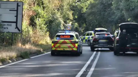 Numerous police cars are at the scene with lights flashing and boots open. They are across both lanes and no traffic can get though.