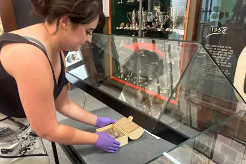 BBC Wales Wearing protective purple rubber gloves, the Curator, wearing a cream and black skirt and black sleeveless top, uses a red rubber sucker to open a hinged display case in order to avoid transferring fingerprints onto the glass. 