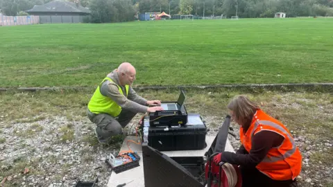 Mining Remediation Authority A woman and a man crouching at the side of a field. Play equipment can be seen in the distance. The man is using a laptop and is wearing a yellow hi-vis top. The woman is closer to the camera, on the right, and wears an orange hi-vis vest and holding some wire.