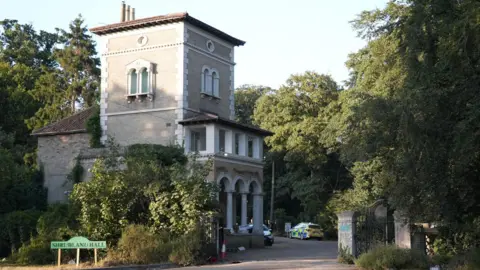 Shaun Whitmore/BBC A gatehouse built over three storeys on the left of an open gate with police cars parked up in the driveway. There are lots of big trees. In front of the gatehouse is a sign saying Shrubland Hall.