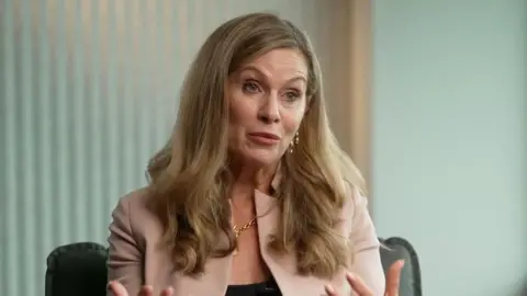 A blonde woman speaks to a camera, wearing a light pink jacket and gold jewelry.