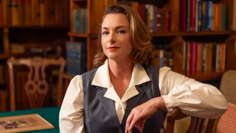 Lillie Sherry Emma is dressed in costume as du Maurier, in a white shirt and waistcoat and smiles out from the photo. She is sat surrounded by old books in a library.