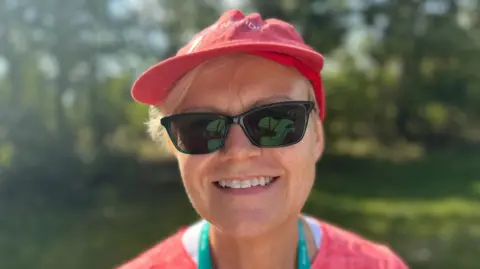 Elliot Deady/BBC A smiling Anne Mills wearing a hat and sunglasses looks straight at the camera.