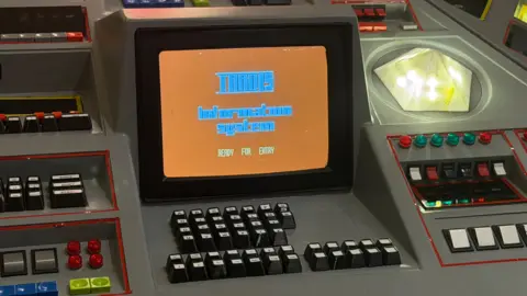 A close-up of a remade version of the buttons and screen of the 1983 TARDIS Console Room. thick black button with white letters on them protrude out of a grey surface. Red, green and blue buttons can be seen around it along with a lit-up diamond-shaped button. In the middle is an orange screen that says 'Tardis Information system, ready for entry'.