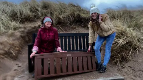 BBC Two women hold up the backrest of the newly-rediscovered memorial bench while sat on another bench on a beach. Marram grass can be seen all around them blowing in the wind. 