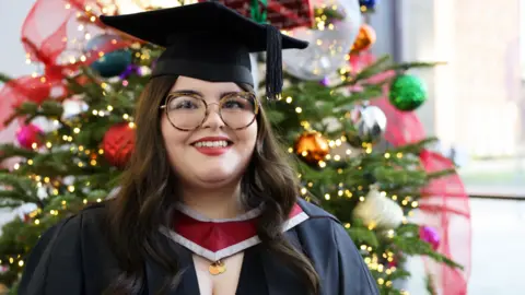 Swansea University Ayla Jones, a white young woman with long brown slightly wavy hair. She has brown eyes and wears large tortoiseshell-framed glasses. She has dark red lipstick on and is dressed in a graduate's cap and gown, with a red and grey-edged piece at the front of her neck. She is standing in front of an out of focus brightly decorated Christmas tree. She is smiling and looking slightly to the right of the camera.