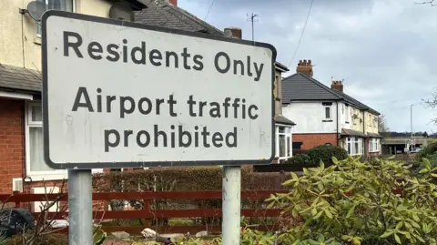 A road sign reads "Residents Only Airport traffic prohibited". Behind is a row of houses.