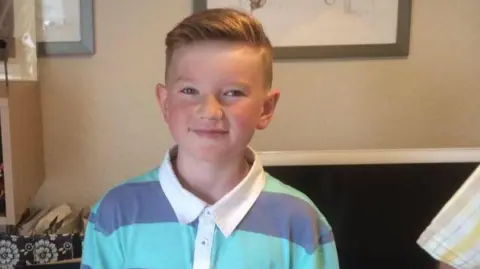11-year-old Alex standing in the foreground facing the camera, wearing a striped polo shirt with a white collar. Behind, a desk or table holds a computer monitor and a patterned storage box with papers. Framed pictures hang on the wall above. On the right edge, part of another person’s shirt sleeve is visible. The indoor setting is softly lit with neutral-coloured walls.
