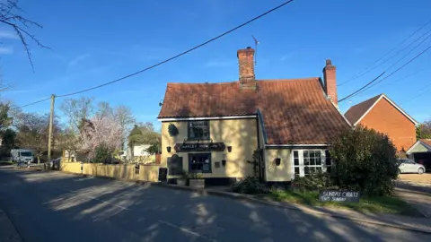Alice Cunningham/BBC A general view of the Barley Mow Inn pub which sits next to a road in a village. The pub is painted yellow with its name painted on a black background.