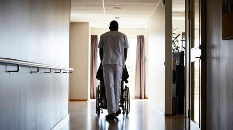 A healthcare worker pushes a wheelchair along a corridor
