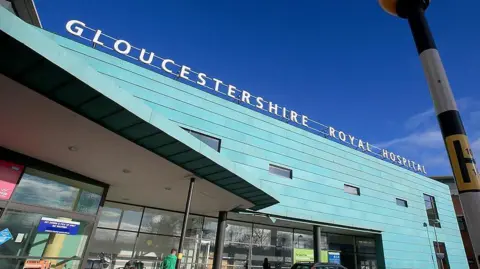 The outside of Gloucestershire Royal Hospital, taken from the road. The entrance to the building is clad in blue/green panels, and the name of the hospital is in large white letters above the building. 
