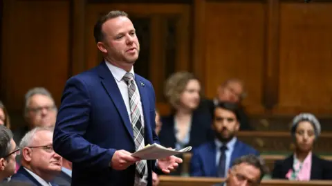 House of Commons/Handout via REUTERS Member of Parliament Ben Maguire speaks during Prime Minister's Questions at the House of Commons in London. He is standing up in the house and is wearing a blue suit and tie. He is holding documents in his hand. People are sitting in benches around him and appear to be listening intently. 