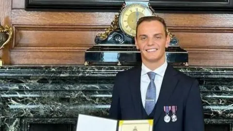 Ben Hawkins standing in a suit wearing two medals on his chest. There is a grand marble fire place behind him and a wood panel wall.