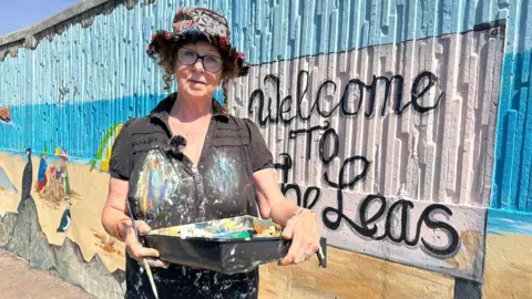John Nurden A woman wearing a hat and painting tools. Her shirt is covered with paint. She is standing in front of a seafront mural.