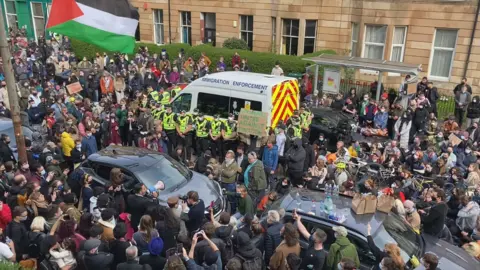 Dozens of people surround an immigration enforcement van on a city street. Several police officers stand in front of the van. 