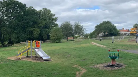 A slide and other play equipment at the play area in Middle Herrington. There are trees and houses across the road in the distance.