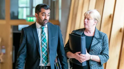 PA Media Humza Yousaf in dark suit and tartan tie walks alongside robison carrying a folder