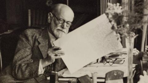 Black and white image of Sigmund Freud sat at a desk looking through papers