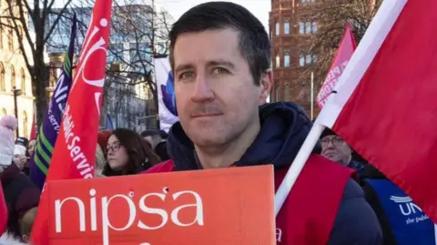 Ryan Wilson with short, dark hair looks into the camera at a Nipsa picket line. He is holding a red Nipsa sign and a red flag. He is surrounded by people who are also holding red and blue Nipsa flags.