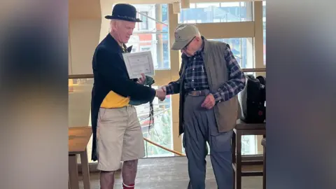 Dull Men's Club A man in a yellow t-shirt, a black bowler hat, a black blazer and beige shorts holds a certificate and shakes hands with a man wearing high-waisted trousers, a navy and pink squared shirt, a green gilet, and a beige cap. 