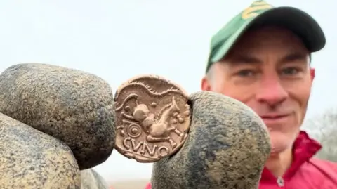 Matt Bell holding up an Iron Age gold coin in his gloved finger and thumb. The coin is in close-up, showing a leaping horse and the letters CVNO. Matt is in the background, a bit blurry, wearing a baseball cap and a red zipped jacket. 