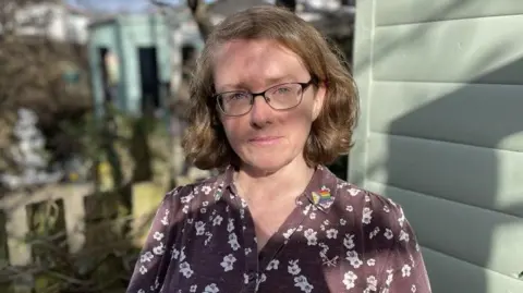 Angela Barron has small glasses and brown, side-parted medium length hair. She is wearing a brown blouse with white flowers on. She is standing beside a pale green garden shed and there are shadows from branches covering her face.