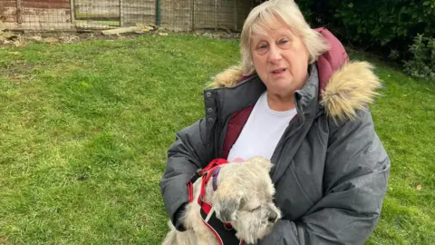 Grace Wood/BBC A woman with a blonde bob and fringe wearing a grey puffer coat and white T shirt holds a small grey and yellow dog.