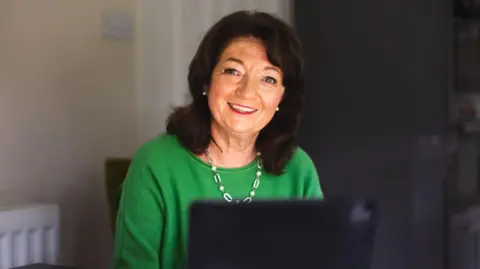 Jersey Brain Tumour Charity Sue smiles at the camera as a computer screen in front of her lights up her face