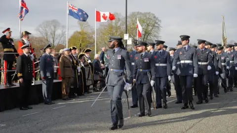 Dozens of people in navy uniforms with armed forces hats and badges are marching in formation along a road. They are looking to the right, towards the Mayor in full red regalia, as well as various other soldiers and personnel, who stand near a raised stage. Great Britain, Canadian and New Zealand flags are flying in the background.