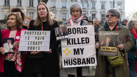 Wiktor Szymanowicz/Future Publishing via Getty Images Members of the Covid-19 Bereaved Families for Justice hold placards - one reads "Boris killed my husband" and another "Partygate is your legacy Boris" - and pictures of loved ones who died during the pandemic as they gather outside Dorland House during the final hearings of the COVID inquiry in London