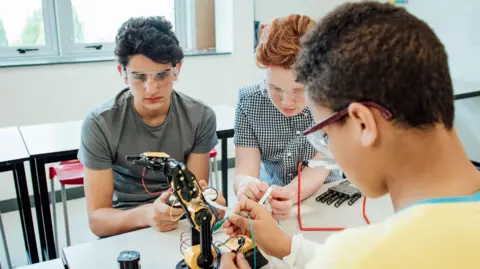 Three students wearing safety goggles work together on a robotic arm in a classroom or laboratory, connecting wires and examining components at a workbench. Tables, chairs and large windows are visible in the bright background.