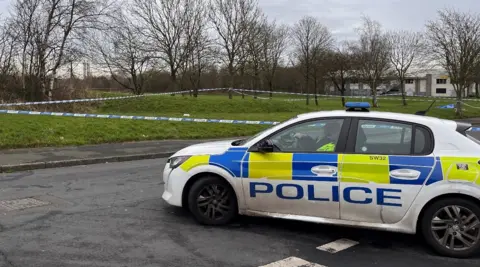 Police tape around a section of grass in the background. A police car is at a junction in the foreground.