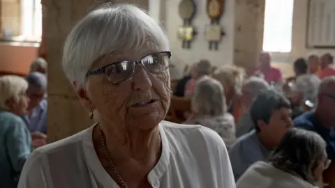 BBC/Crispin Rolfe Pam has short grey hair and black-framed specs on her face. She is wearing a white blouse and is standing in a room full of people sitting at tables.